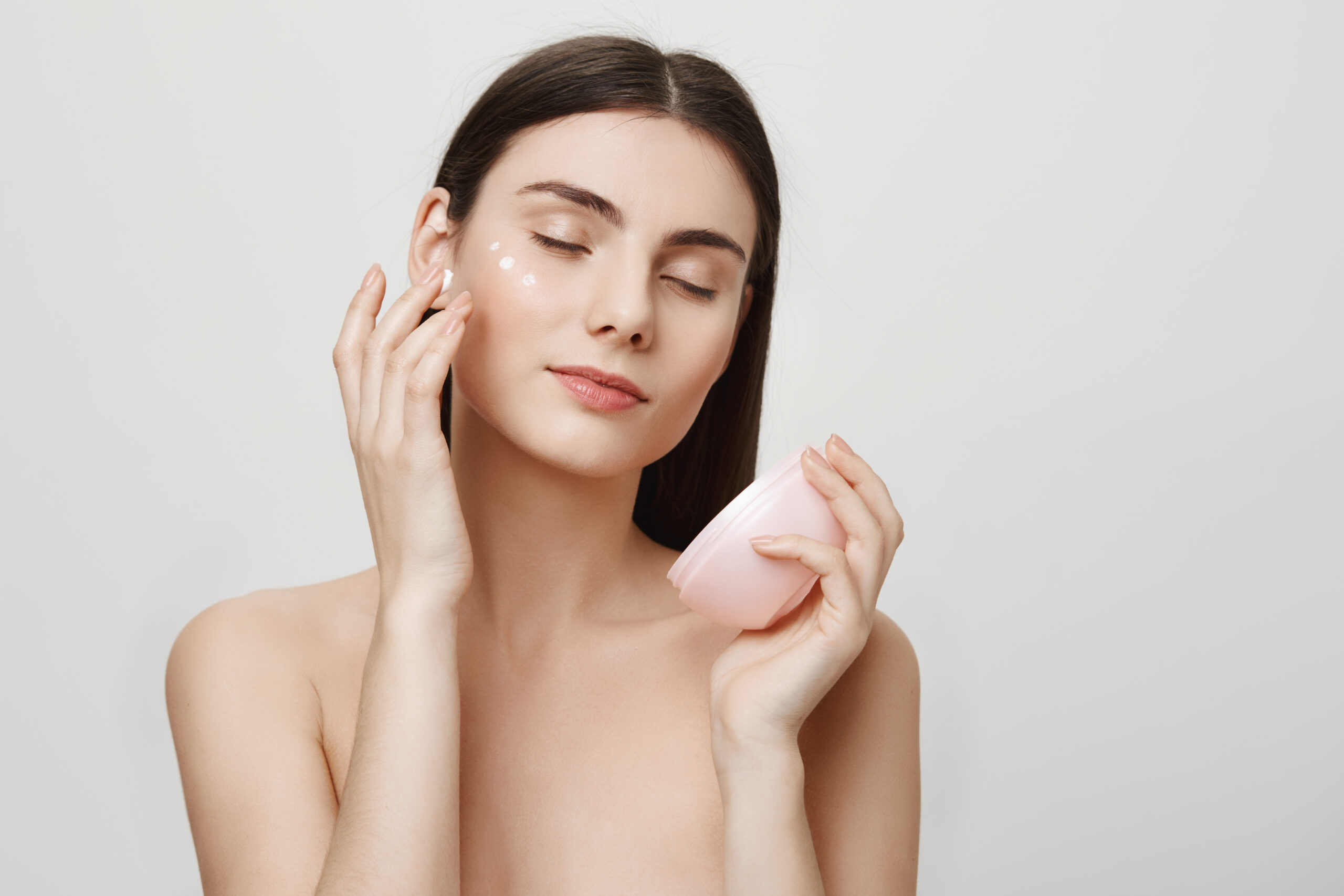 Beauty and skincare concept. Studio portrait of gentle good-looking european woman rubbing in facial cream, enjoying doing cosmetological procedures while standing over gray background.